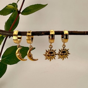 Peut inclure: Quatre boucles d'oreilles dorées. Deux boucles d'oreilles présentent des breloques en forme de croissant de lune, et les deux autres ont des breloques en forme de soleil. Les boucles d'oreilles sont présentées sur une branche brune avec des feuilles vertes en arrière-plan.