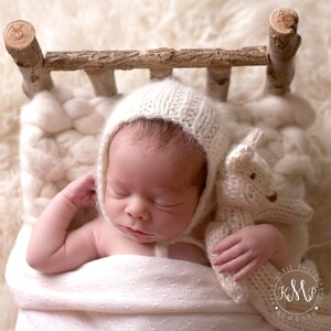 May include: A newborn baby wearing a white knitted hat and sleeping in a wooden crib with a white knitted blanket and a white knitted stuffed animal.