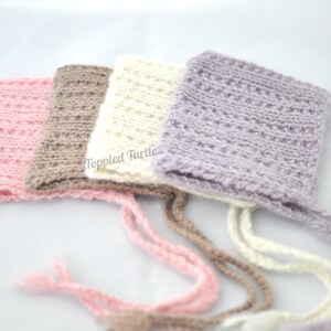 May include: Four crocheted baby bonnets in pink, brown, white, and purple with matching ties. The bonnets are made with a textured stitch and have a simple, classic design.