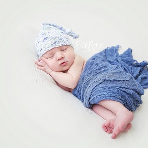 Puede incluir: Un bebé recién nacido con un gorro de punto blanco y azul duerme en un arrullo de tela azul. El bebé está acostado de lado con la cabeza apoyada en el brazo.
