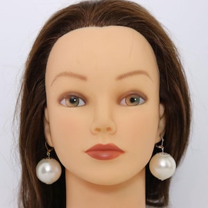 May include: A pair of large, round, white pearl earrings with gold-colored hooks. The earrings are displayed on a mannequin head with brown hair, fair skin, and brown and green eyes. The earrings are a classic jewelry accessory.