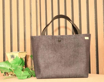 May include: A dark gray tote bag with black handles and a button closure. The bag is rectangular and has a small tag on the side. A small decorative pot and green leaves are in the background.