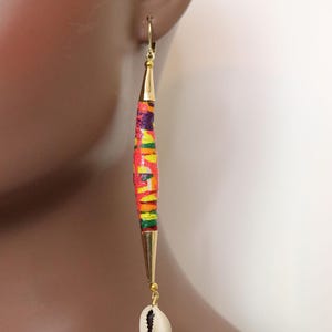 May include: Long, colorful dangle earrings with a cylindrical design. The earrings feature a vibrant pattern of red, yellow, green, and purple. Gold-colored metal accents and a small white seashell complete the design. These earrings are a unique accessory.