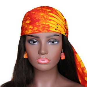 May include: A mannequin with long black hair wearing a bright orange and yellow bandana with black accents. The bandana is tied around the head and flows down the back. The mannequin is wearing orange earrings.