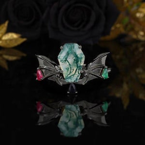 May include: A gothic-style ring with a central, coffin-shaped green gemstone. The ring features black metal wings on either side, with a red and green gemstone accent. The ring is displayed on a reflective black surface.