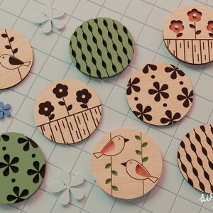 May include: A collection of round wooden coasters with various designs. Some feature birds, flowers, and geometric patterns in black, green, and pink. The coasters are arranged on a light blue tiled surface, with decorative flower accents.