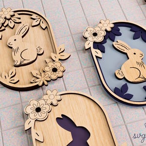 Op de afbeelding: Houten paasdecoraties met konijntjes en bloemmotieven. De konijntjes zijn lichtbruin met donkerbruine contouren en de bloemen zijn wit en donkerbruin. De achtergrond is lichtblauw en paars.