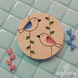 May include: A wooden round with two birds, one with blue and the other with pink accents, perched on green-leafed vines. Two additional leaf sprigs, one pink and one blue, are placed nearby. The background is a light blue tiled surface.