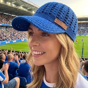 May include: A blue crochet baseball cap with a brown leather patch on the side. The cap has a visor and an open weave design. The person is wearing a blue and white shirt and is at a sporting event.