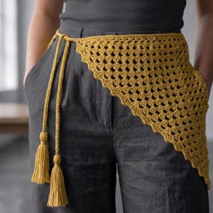 May include: Mustard yellow crochet belt with a triangular design. The belt has a long, braided tie with tassels at the ends. The belt is worn over gray pants and a gray top.