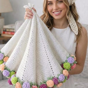 May include: A cream-colored, crocheted baby blanket with a floral border. The blanket is held up, showcasing the intricate stitchwork and the colorful, handmade flowers in shades of pink, peach, purple, and green. The blanket is approximately 36 inches in diameter.