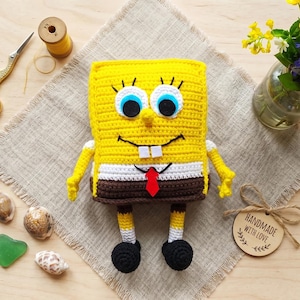 May include: A handmade, crocheted SpongeBob SquarePants plush toy. The toy is yellow with blue eyes, a black smile, and a red tie. It has white arms and legs with black shoes. A wooden tag reads "HANDMADE WITH LOVE."
