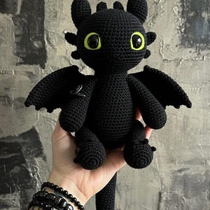 Puede incluir: Un peluche de dragón negro tejido a ganchillo con grandes ojos amarillos y pequeñas alas. El dragón tiene una cola larga y está hecho de un hilo suave y texturizado. El artículo está hecho a mano.