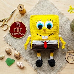 May include: A handmade crochet figure of SpongeBob SquarePants, featuring yellow body, blue eyes, and a red tie. The figure is placed on a textured surface with seashells and a "PDF ESPAÑOL" button.