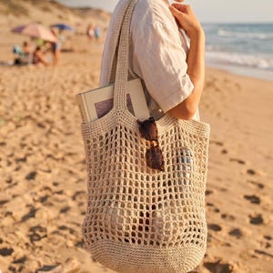 May include: A beige, open-weave crochet beach tote bag with a book and sunglasses inside. The bag is held by a person on a sandy beach. Text on the bag reads "PDF Pattern Boho Beach Tote" and "Eco-friendly Cotton".