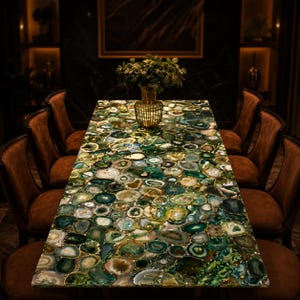 Può includere: Un tavolo da pranzo rettangolare con piano in pietra di agata verde. Il tavolo è circondato da otto sedie imbottite marroni. Un vaso d'oro con fogliame verde è al centro del tavolo. La stanza ha boiserie scure e un grande dipinto.