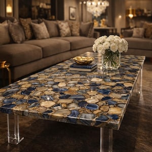 Pode incluir: Uma mesa de centro retangular com um tampo em mosaico com pedras azuis, bege e douradas. A mesa tem pernas de acrílico transparente. Um vaso com rosas brancas, uma tigela dourada e livros estão sobre a mesa. Numa sala de estar.