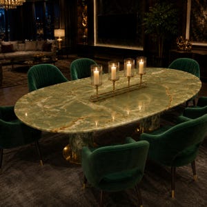 Op de afbeelding: Een ovale groene marmeren eettafel met gouden accenten, omringd door zes groene fluwelen stoelen. Een gouden kandelaar met vier brandende kaarsen staat op de tafel. De kamer heeft een donkere, luxueuze uitstraling.
