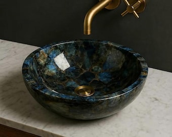 Puede incluir: Un lavabo de sobreponer redondo fabricado en piedra pulida de tonos azules y oscuros con vetas naturales. El lavabo cuenta con un desagüe central de latón y se combina con un grifo de pared de latón y un mando de cruz en una pared oscura.