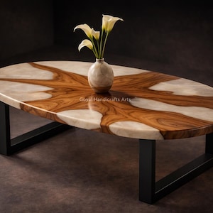 Könnte beinhalten: Ovaler Couchtisch mit einer Tischplatte aus Holz und Harz. Die Tischplatte hat ein einzigartiges Muster aus hellen und dunklen Holztönen mit einer weißen Harzfüllung. Der Tisch hat schwarze Metallbeine und eine kleine Vase mit weißen Blumen.