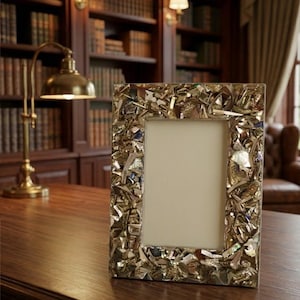 Puede incluir: Un marco de fotos rectangular con un borde de mosaico hecho de varias piezas metálicas e iridiscentes. El marco tiene un interior de color neutro y se encuentra sobre una superficie de madera oscura, con una biblioteca y una lámpara en el fondo.