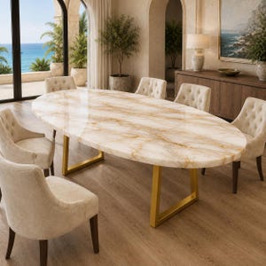 May include: An oval dining table with a polished white and gold marble-patterned top supported by two angular gold metal legs. The table is surrounded by six beige upholstered chairs featuring button-tufted backrests and dark wooden legs.