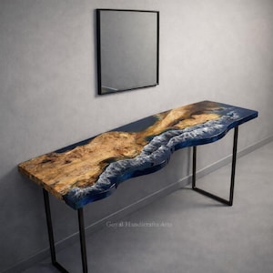 May include: A long, rectangular console table with a live edge design. The tabletop features a wood grain pattern with a blue resin river and white wave details. The table is supported by black metal legs. A black framed mirror hangs above.