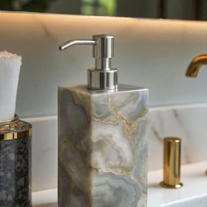 Puede incluir: Un dispensador de jabón rectangular con un patrón similar al mármol en tonos grises, blancos y dorados. Tiene una bomba plateada y se exhibe sobre una encimera blanca. El logotipo "GoyalHandicraftsArts" es visible.