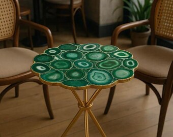 May include: A decorative side table with a unique design. The tabletop is made of green agate slices with a gold trim. The table has a gold metal tripod base. The table is in a room with wooden chairs and a wooden floor.