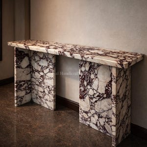 Puede incluir: Una consola rectangular de mármol con un patrón blanco y burdeos. La mesa tiene una parte superior gruesa y dos patas de soporte. La mesa está hecha de mármol y tiene un acabado pulido. La mesa está en una habitación con un espejo y un suelo marrón oscuro.