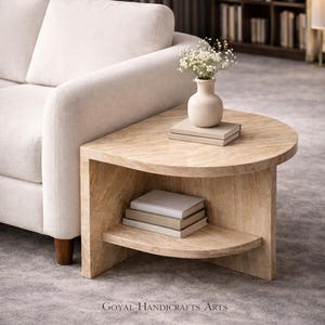 以下が含まれることがあります： 明るいベージュ色の半円形のサイドテーブル。下段の棚には本が積まれています。上には白い花が入った小さな花瓶があります。テーブルはクリーム色のソファの隣にあります。「GOYAL HANDICRAFTS ARTS」の文字が見えます。