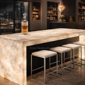 Könnte beinhalten: Eine hinterleuchtete Bar mit einer hellen Arbeitsplatte und vier quadratischen Hockern mit silbernen Beinen. Eine Kristallkaraffe mit bernsteinfarbener Flüssigkeit steht auf der Bar. Die Bar befindet sich in einer modernen Umgebung mit Blick auf die Skyline.