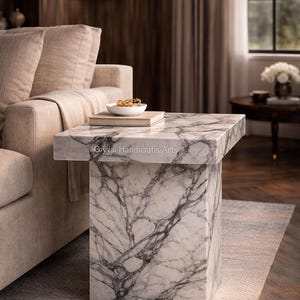 May include: A marble side table with a rectangular top and a square base. The table features a white and gray veined pattern. A bowl and a book rest on the table's surface. The table is next to a beige sofa.