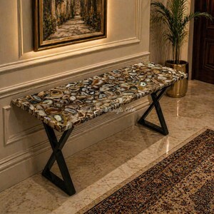May include: A rectangular bench with a mosaic agate stone top, featuring brown, white, and gray patterns. The bench has black metal X-shaped legs. The bench is placed in a room with a painting and a potted plant.