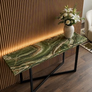 Peut inclure: Une console en onyx vert avec une base en métal noir. La table présente un dessus en pierre naturelle avec des motifs tourbillonnants de vert, de brun et de blanc. Un vase de fleurs blanches est posé sur la table. La table mesure 152 cm de long.