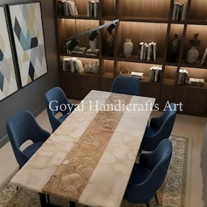 May include: A rectangular dining table with a marble top and a central brown stripe, surrounded by white marble. The table is set with six blue velvet chairs. The text "Goyal Handicrafts Art" is visible on the table. The room has a bookshelf and artwork.