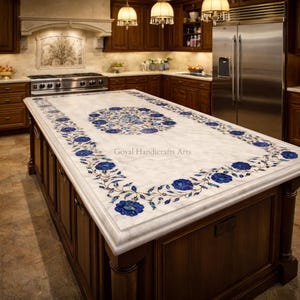 May include: A white marble kitchen island countertop with intricate blue floral inlay designs. The island is made of dark wood and features the text "Goyal Handicrafts Arts". The countertop is a focal point in a well-lit kitchen.