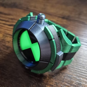May include: A green, black, and silver Omnitrix watch prop. The watch has a circular face with a green and black symbol, framed by a silver bezel. The strap is green with black details. A silver button is on top.