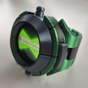 May include: A green and grey toy watch with the text "02 15" in white on a green screen. The watch has a round face and a segmented band. The watch is made of plastic and has a button on the top.
