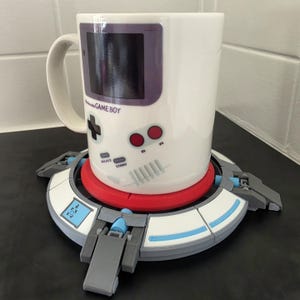 May include: A white ceramic mug featuring a graphic of a classic handheld gaming console with the text Nintendo GAME BOY, resting on a circular, multi-coloured plastic base with grey, white, red, and light blue segments and mechanical details.