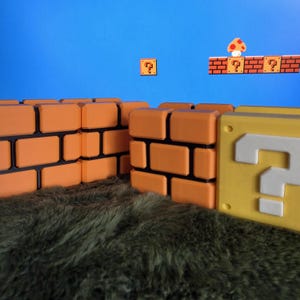 May include: A collection of orange brick blocks and a yellow question mark block arranged on a textured green surface. In the background, a blue screen displays a digital scene featuring floating brick platforms, question blocks, and a mushroom.