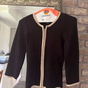 May include: Black zip-up cardigan with cream-colored trim and a matching cream-colored lining around the neckline. The long-sleeved sweater is hanging on a Tide Cleaners hanger.