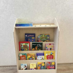 May include: A light-coloured wooden bookshelf filled with colourful children's books. The bookshelf has multiple shelves and a slanted design, with books of various sizes and colours displayed. A stack of blue books sits on the top shelf.