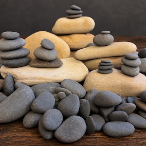 May include: A collection of smooth, rounded river stones in shades of dark gray and light tan. Several stones are stacked into small, balanced towers of varying heights, while others are scattered loosely across the foreground surface.