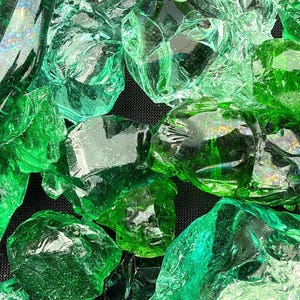 May include: Close-up of a collection of irregularly shaped, translucent green glass pieces. The glass shards vary in size and shape, with a range of light and dark green hues. The pieces are scattered, creating a textured, sparkling effect.