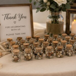 May include: A collection of small, clear glass jars with cork stoppers, each filled with tiny, multi-colored pebbles. Beside them stands a wooden-framed sign with the text: Thank You, FOR CELEBRATING WITH US, PLEASE TAKE ONE.
