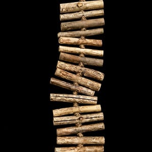 May include: A vertical hanging decoration composed of multiple light brown wooden sticks strung together with natural fiber rope. The structure features a top loop for hanging, a smooth wooden bead at each end, and a single flat stone at the bottom.