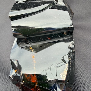 May include: A large, irregularly shaped piece of obsidian. The polished surface reflects light, showing a range of colors from deep black to reddish-brown. The edges are jagged, revealing the natural fracture of the volcanic glass.