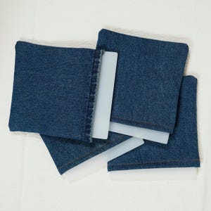 May include: Four blue denim coasters with white plastic inserts are arranged on a white surface. The coasters are square and appear to be made from recycled denim. The white inserts are rectangular and provide a contrasting element.