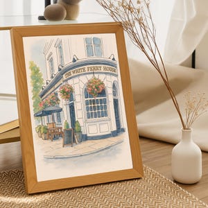 May include: A framed watercolor print of a pub, "The White Ferry House", with a light brown wooden frame. The building is white with blue accents, floral arrangements, and a black awning. A small chalkboard stands in front.